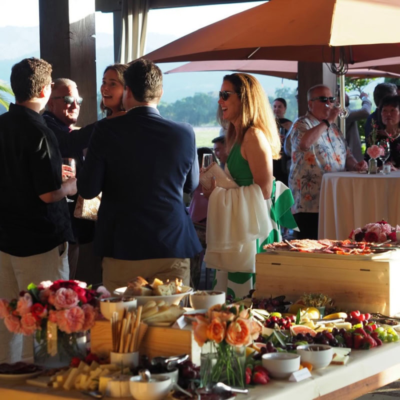 A group of guests enjoying drinks and conversation at a holiday celebration at Round Pond Estate, with a buffet table of food.