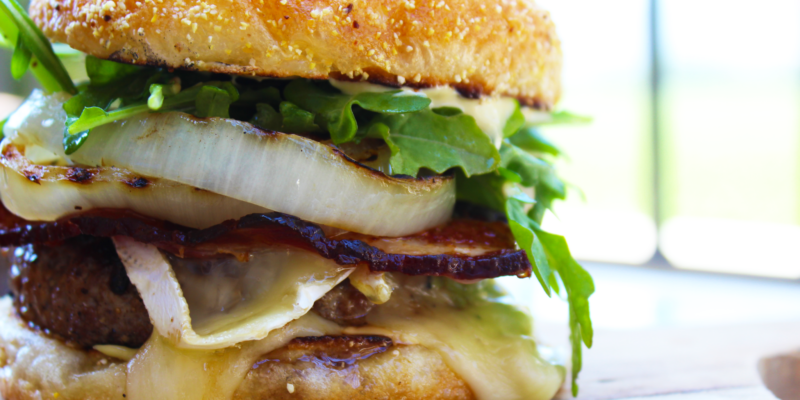 Close-up of a gourmet brisket burger topped with Brie cheese, applewood-smoked bacon, fresh greens, and caramelized onions on.