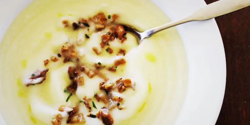 Delicious creamy cauliflower soup garnished with crispy bacon bits and fresh herbs, served in a white bowl with a spoon.