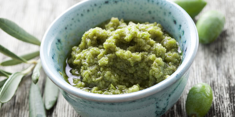 Close-up of green olive tapenade in a ceramic bowl, with fresh olives and herbs in the background.