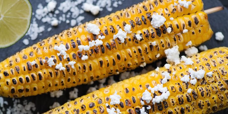 Close-up of grilled corn on skewers topped with chili-lime seasoning and crumbled cheese, on a dark background.