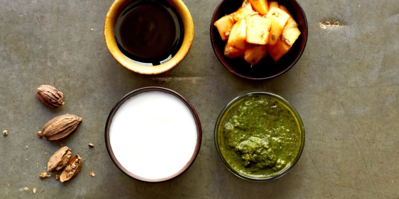 Close-up of herb chutney, yogurt, and spices on a gray surface, with a cup of dark liquid and chopped herbs.