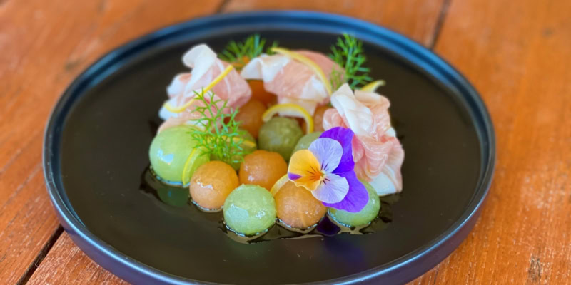 Delicious melon and prosciutto salad garnished with edible flowers and fresh herbs on a black plate.