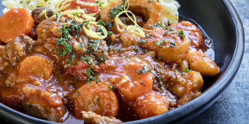 A close-up of tender veal osso buco with carrots, herbs, and rich sauce in a black dish.