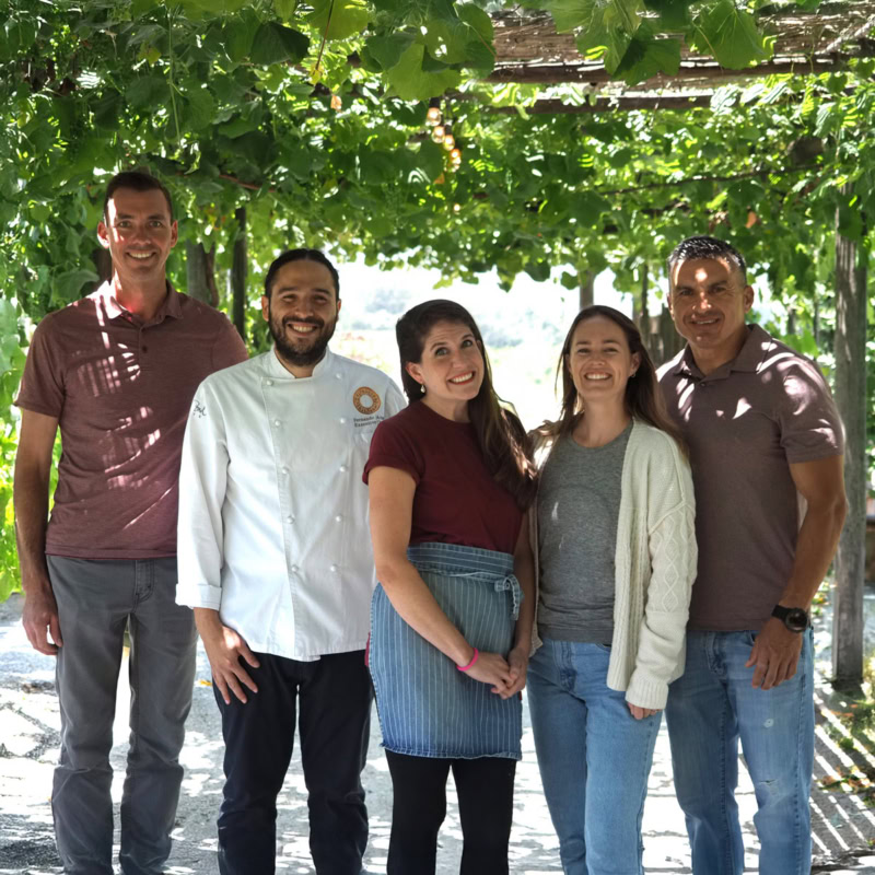 Chef and team members smiling under lush grapevine canopy.