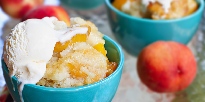 Delicious peach cobbler topped with vanilla ice cream, served in a turquoise bowl with fresh peaches nearby.