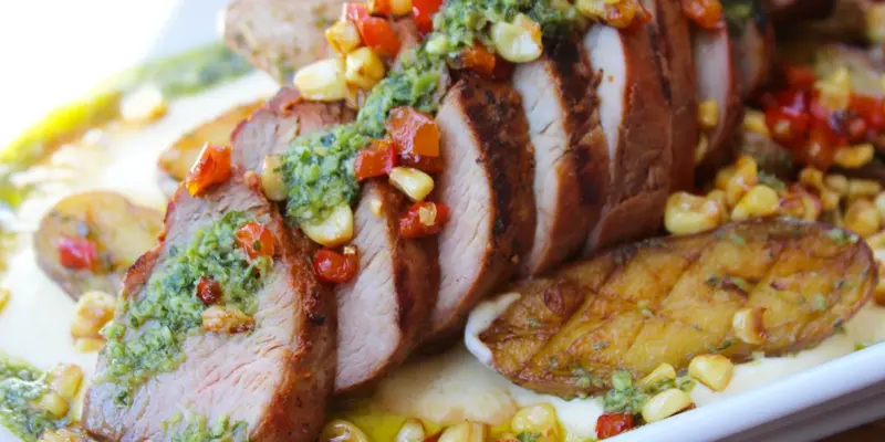 Close-up of sliced pork tenderloin topped with herb verde sauce, served with crispy potatoes and colorful corn salsa.