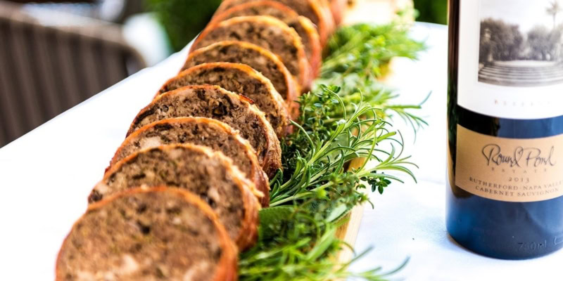 Close-up of sliced meatloaf with herbs and a bottle of red wine on a white plate.