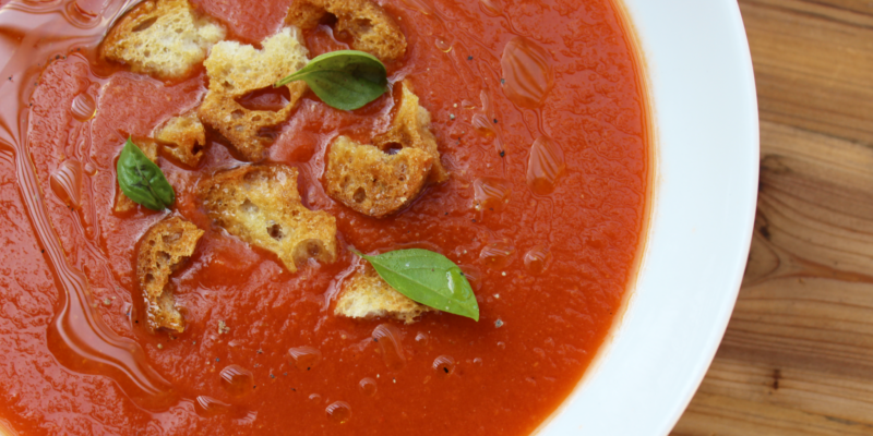 Fresh heirloom tomato and roasted red pepper gazpacho in a white bowl, garnished with croutons and basil leaves.