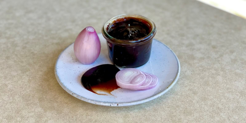 Close-up of a small jar of red wine shallot jam with a shallot and balsamic vinegar on a white plate.