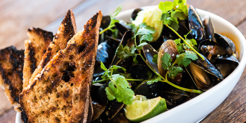 Bowl of steamed mussels garnished with cilantro and lime wedges, served with toasted bread slices on a wooden table.