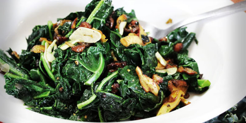 Sautéed kale and mushrooms with crispy bacon pieces, served on a white plate.