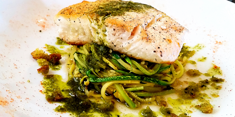 A plate of zucchini noodles topped with a seared halibut fillet, garnished with green herbs and drizzled with a light sauce.