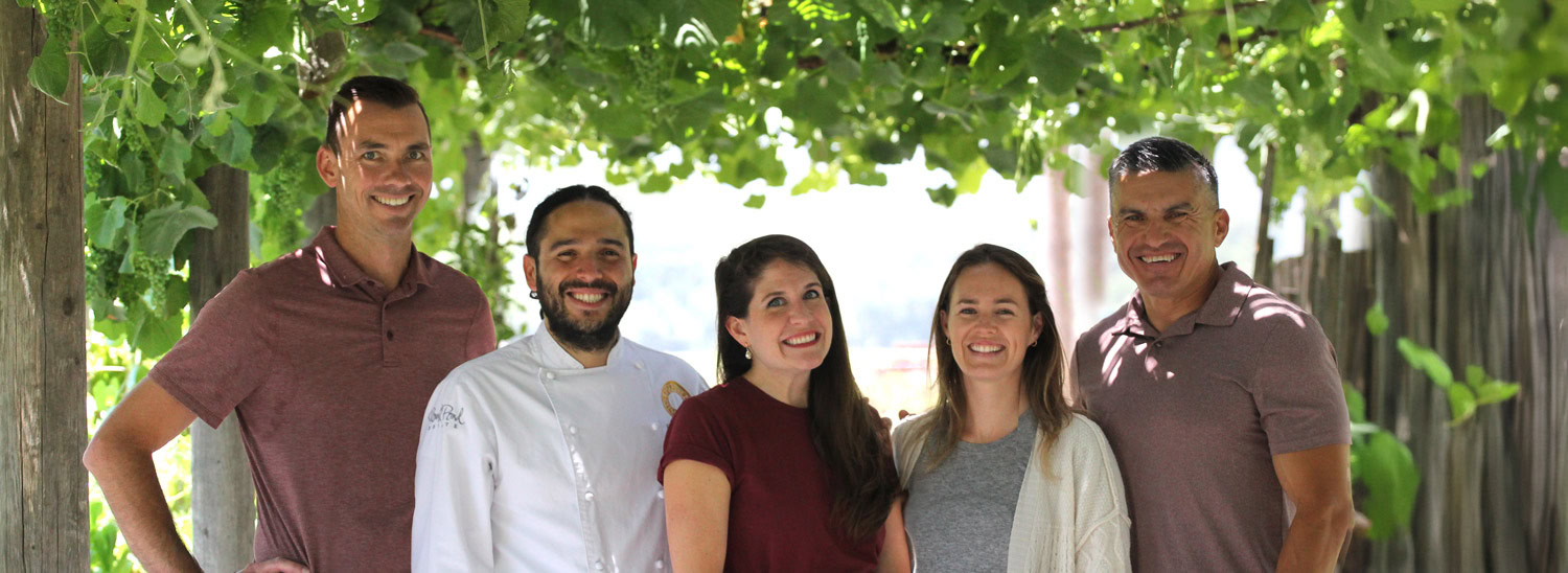 Group of five diverse people smiling outdoors under grapevines at Round Pond Estate.