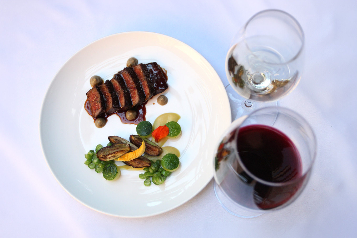 Gourmet steak dish with vegetables and two glasses of red and white wine on a white tablecloth.